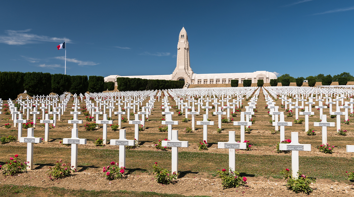 rows of white crosses marking graves stretching over a green hill with a white monument and French flag in the distance